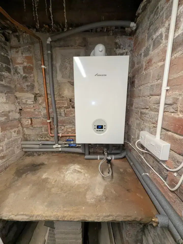 Gas boiler mounted on brick wall with pipes and electrical connections in a utility room.