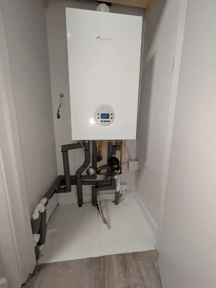Modern white boiler with control panel installed in a small utility room, plumbing pipes visible beneath.