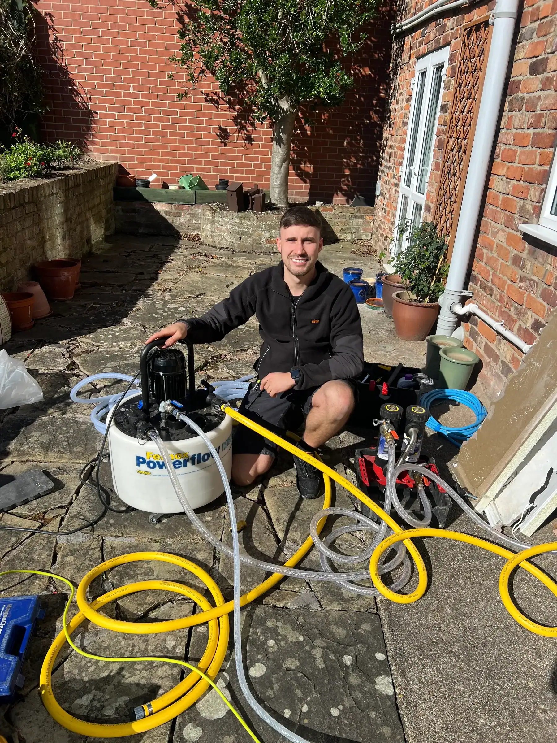 Man working on air conditioning unit outside a brick house in a garden with tools and yellow hoses.