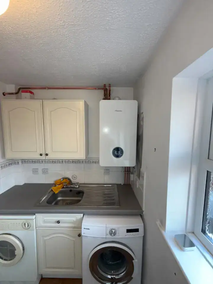 Gas boiler installed above kitchen sink with washing machine and cupboard in a small utility room.